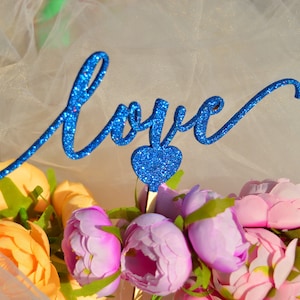 Puede incluir: Un adorno para pastel de purpurina azul con la palabra "love" en letra cursiva y un peque&ntilde;o coraz&oacute;n en el centro. El adorno est&aacute; sobre un ramo de flores rosas y naranjas.