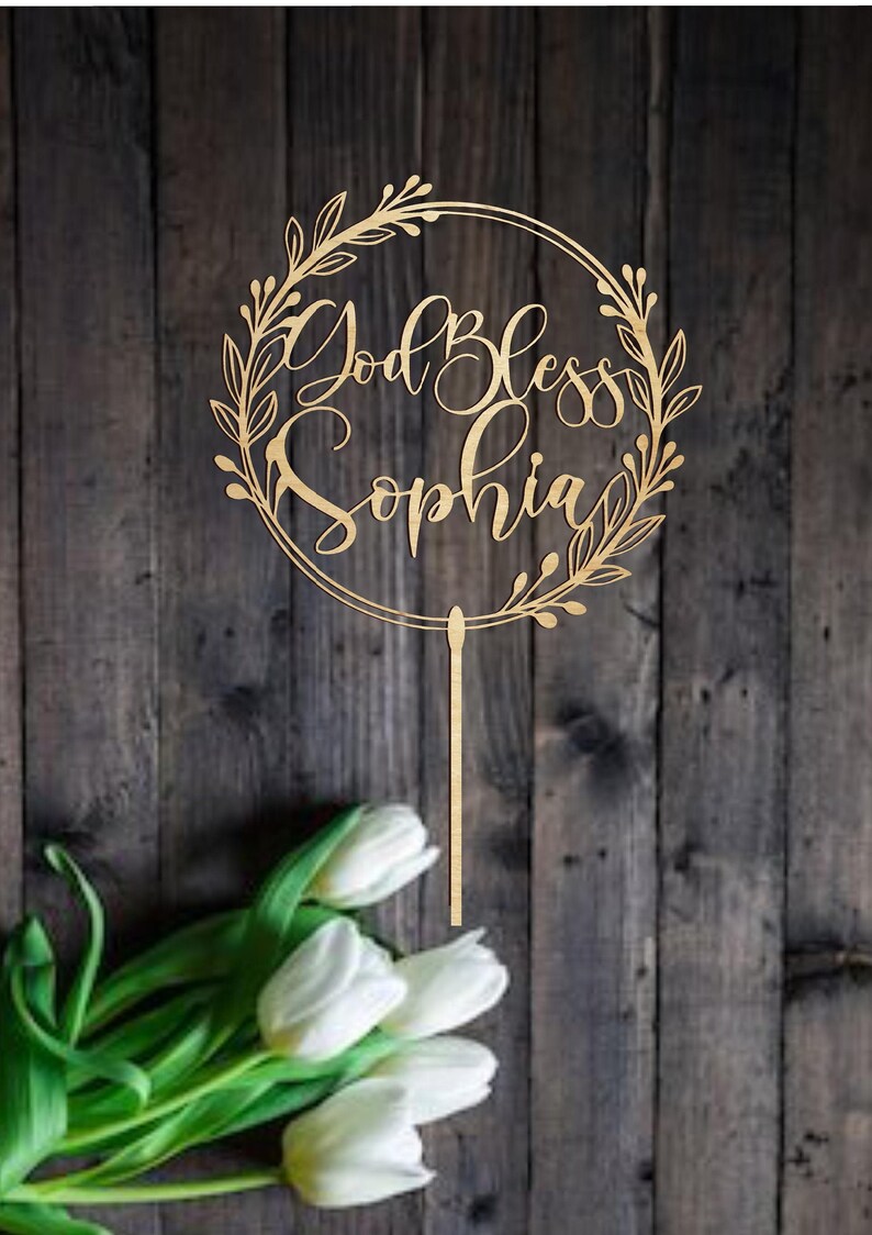 Puede incluir: Un adorno para pastel de madera con un dise&ntilde;o de corona circular. La corona est&aacute; hecha de hojas y tiene las palabras "God Bless Sophia" escritas en su interior. El adorno est&aacute; en un palo de madera.