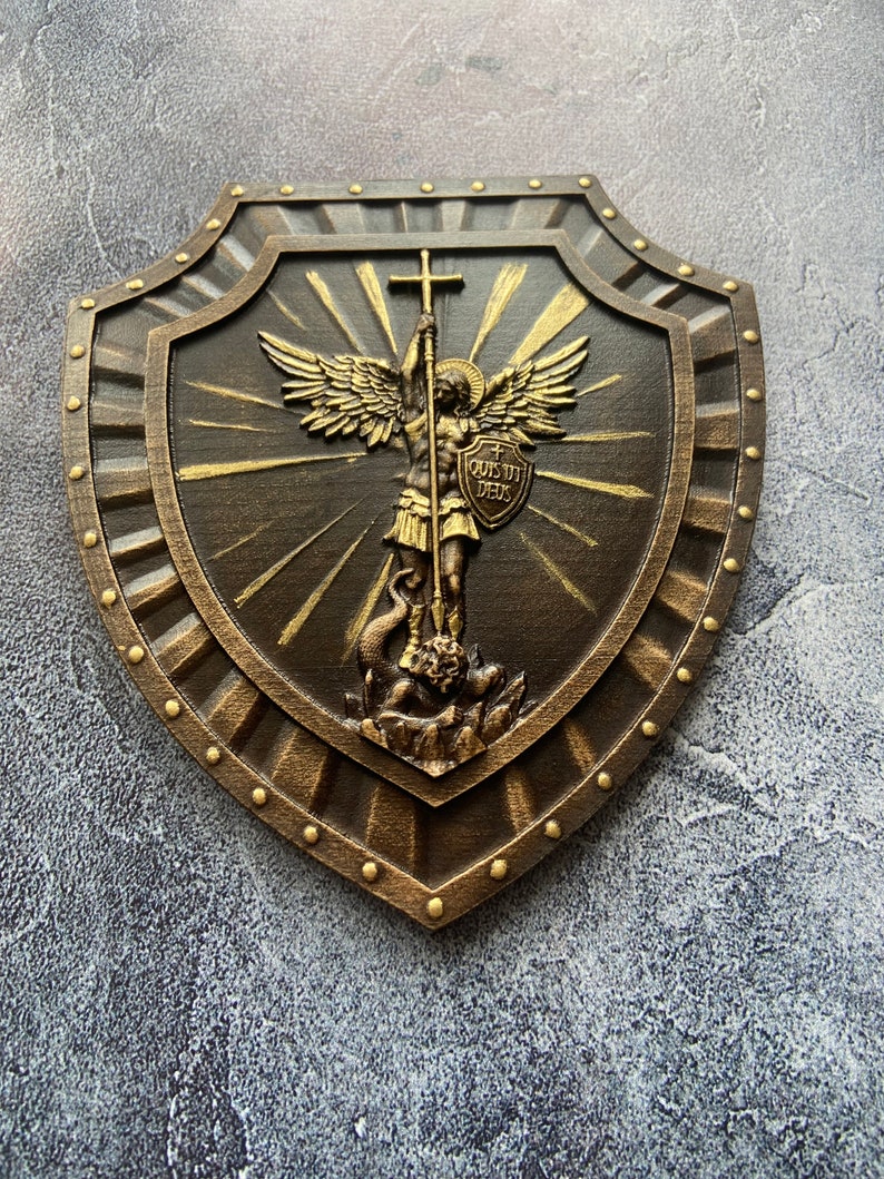 May include: A brown wooden shield with a gold-painted design of an angel holding a cross and a shield with the text "QUIS UT DEUS".