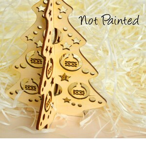 Puede incluir: Adorno de &aacute;rbol de Navidad de madera sin pintar con un dise&ntilde;o recortado de estrellas y bolas. El &aacute;rbol est&aacute; sobre un lecho de papel blanco triturado. "Not Painted" est&aacute; escrito en texto negro sobre el papel blanco.