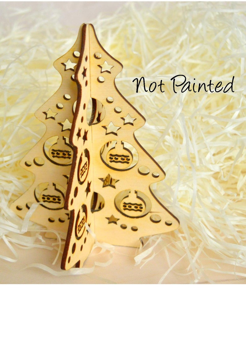 Puede incluir: Adorno de &aacute;rbol de Navidad de madera sin pintar con un dise&ntilde;o recortado de estrellas y bolas. El &aacute;rbol est&aacute; sobre un lecho de papel blanco triturado. "Not Painted" est&aacute; escrito en texto negro sobre el papel blanco.