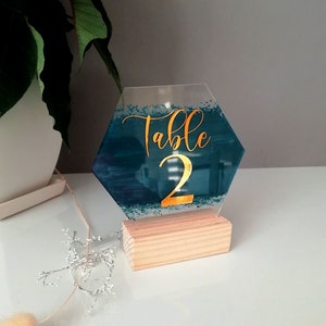 Puede incluir: Un número de mesa hexagonal de acrílico azul turquesa con letras doradas de purpurina que dice "Mesa 2" sobre una base de madera.
