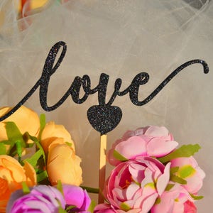 Puede incluir: Un letrero de 'love' de purpurina negra con un coraz&oacute;n, rodeado de flores rosas y amarillas sobre un fondo de tul blanco.