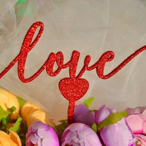 Puede incluir: Un letrero de "Love" rojo con purpurina con un coraz&oacute;n rojo con purpurina, sobre un fondo de tul blanco con un ramo de flores amarillas y moradas.
