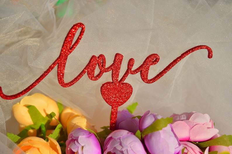 Puede incluir: Un letrero de "Love" rojo con purpurina con un coraz&oacute;n rojo con purpurina, sobre un fondo de tul blanco con un ramo de flores amarillas y moradas.