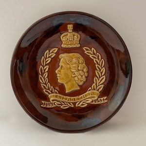 May include: A brown ceramic plate with a raised image of Queen Elizabeth II in profile, wearing a crown and surrounded by a laurel wreath. The plate is inscribed with "June Elizabeth R 1953".
