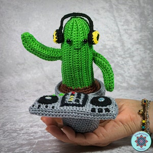 Puede incluir: Un cactus de crochet verde con auriculares y sosteniendo un tocadiscos de crochet gris. El cactus está en una maceta gris.
