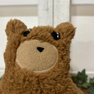 May include: A brown plush bear toy featuring a fuzzy, textured exterior, a smooth tan circular muzzle, two small black embroidered eyes, and a small black triangular nose. The bear has rounded ears and a soft, stitched construction throughout.