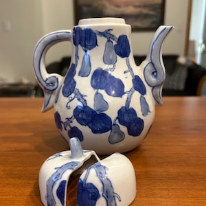 May include: A white ceramic teapot featuring a blue hand-painted pattern of pears and leafy branches. The teapot has a curved handle and spout. In front of the teapot lies a broken ceramic lid, split into two pieces with matching blue designs.