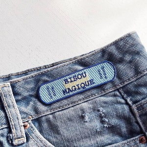 Peut inclure: Une poche en jean bleu avec un patch brodé bleu et jaune qui dit "Bisou Magique".