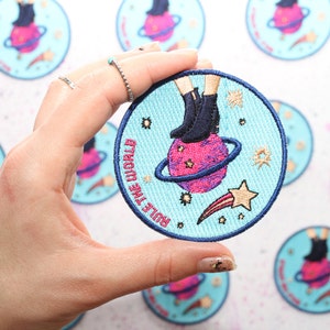 Puede incluir: Un parche bordado azul y blanco con una figura de dibujos animados de pie en un planeta con el texto "Rule the World".