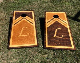 May include: Two wooden cornhole boards with a brown stain and a light wood inlay. The boards have a chevron pattern and the letter "L" in a light color.