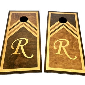 May include: Two wooden cornhole boards with a brown stain and a light wood border. The boards have a chevron design and the letter "R" in gold lettering.