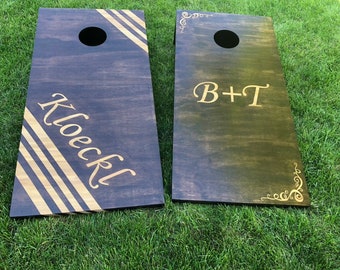 May include: Two wooden cornhole boards with a dark brown stain. One board has the name "Klockl" in gold lettering with gold stripes. The other board has the initials "B+T" in gold lettering with gold swirls in the corners.