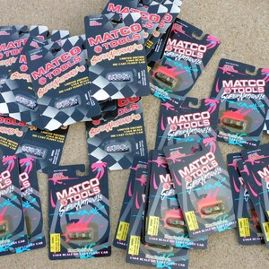 Può includere: Una collezione di modellini di auto da corsa Matco Tools Super Nationals nelle loro confezioni originali. La confezione è nera con accenti rosa e blu e presenta un design a bandiera a scacchi. Ogni auto è in scala 1/64.
