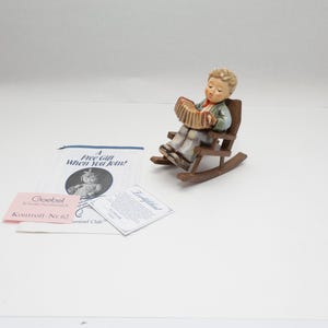 May include: A ceramic figurine of a child with blonde hair sitting in a brown wooden rocking chair and playing a tan accordion. Included are a white paper pamphlet with the text A Free Gift When You Join and a small pink Goebel Kontroll-Nr.62 tag.