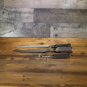 May include: A three-piece carving set comprising a sharpening steel, a carving knife, and a carving fork. The handles are dark brown, and the metal parts feature ornate silver-coloured detailing. The set is presented on a wooden surface.