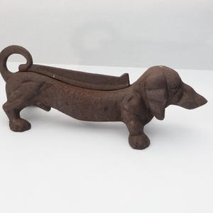 May include: A brown, textured cast iron boot scraper shaped like a dachshund dog. The dog figure is standing with its head facing right, featuring a curled tail and a long, low body design with a flat, elevated ridge along the back for scraping.