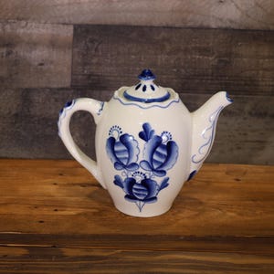 May include: White ceramic teapot with blue floral designs. The teapot features a curved handle, a spout, and a lid with a small knob. The blue floral pattern is repeated on the handle and lid, creating a cohesive design.