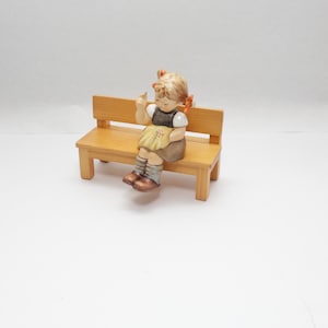 Może przedstawiać: Ceramiczna figurka dziecka z blond kucykami siedzącego na jasnobrązowej drewnianej ławce. Dziecko ma na sobie białą koszulę, ciemną kamizelkę, żółtą spódnicę i brązowe buty, trzymając w jednej dłoni mały przedmiot i patrząc w dół.
