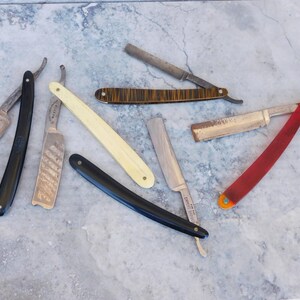 May include: Collection of vintage straight razors with various handle colors, including black, cream, and red. Blades show signs of age and use, with some featuring decorative etching. The razors are arranged on a mottled gray surface.