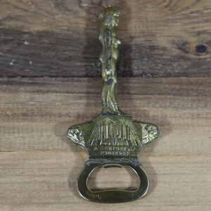 May include: Vintage brass bottle opener with a detailed figure at the top and a decorative base. The base features an architectural design and the text "A Century of Progress". The opener is made of brass and has a circular opening.