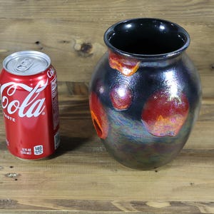 Könnte beinhalten: Eine dekorative, irisierende Vase mit dunklem, glänzendem Finish und roten und orangen Akzenten. Eine rote Coca-Cola-Dose dient zum Größenvergleich. Die Vase hat einen breiten Körper und eine schmale Öffnung.
