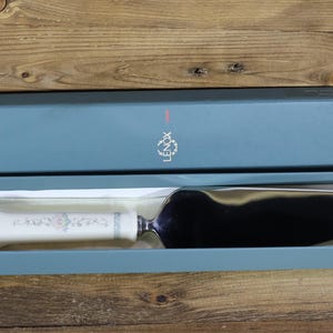 Può includere: Un server per torte con manico a fantasia floreale, presentato in una scatola blu. Il server ha una lama in acciaio inossidabile e un manico bianco con accenti floreali rosa e blu. La scatola ha il logo Lenox.