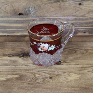 Pode incluir: Uma chávena de chá de vidro transparente com uma pega, com um design floral decorativo e uma faixa castanho-avermelhada com acabamento dourado. A chávena tem uma base facetada e um interior castanho-avermelhado com texto cursivo.