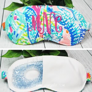 May include: A colorful eye mask with a floral pattern in shades of blue, pink, and green, embroidered with the initials "MNE" in pink. The mask is shown open, revealing a gel bead insert. The website address www.sewgirlie.com is printed on the mask.