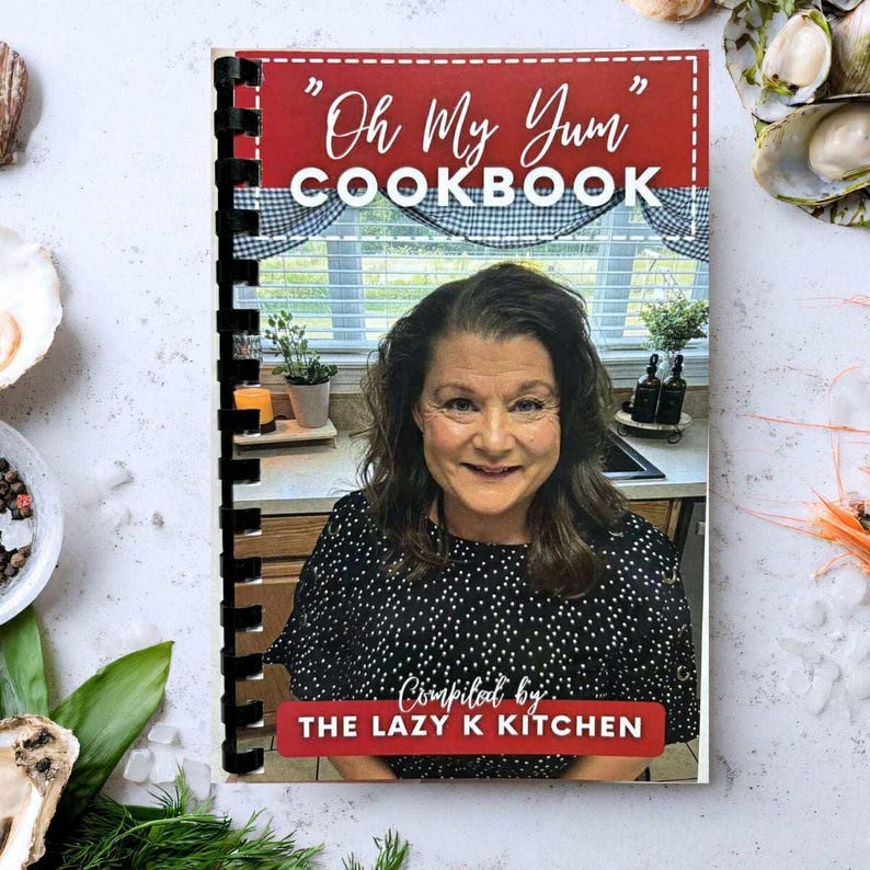 May include: A spiral-bound cookbook titled "Oh My Yum COOKBOOK" with a red banner. The cover features a smiling woman in a black and white polka dot top. The text "Compiled by: THE LAZY K KITCHEN" is also visible.