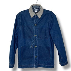 May include: A blue denim jacket with a tan collar and button closure. The jacket has two chest pockets and two lower pockets. The jacket is hanging on a metal hanger against a white background.