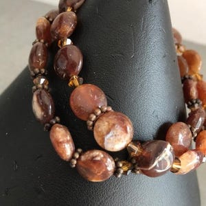 Peut inclure: Gros plan sur un bracelet à deux rangs composé de perles ovales et rondes dans des tons de marron, ambre et crème. Les perles sont enfilées avec de plus petites perles de couleur bronze et des accents dorés. Le bracelet est présenté sur un présentoir noir.