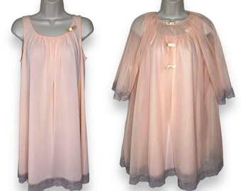 Op de afbeelding: Een lichtroze vintage lingerie set, bestaande uit een mouwloze nachtjapon en een doorschijnende robe. Beide stukken hebben grijze kanten afwerking aan de zoom en manchetten. De nachtjapon heeft een ronde halslijn en een decoratief goudkleurig accent. De robe heeft een ronde kraag en goudkleurige sluitingen.