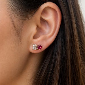 Puede incluir: Pendientes de oro con tachuelas, uno con una piedra transparente rectangular y el otro con una piedra roja en forma de corazón. Los pendientes se llevan en una oreja.