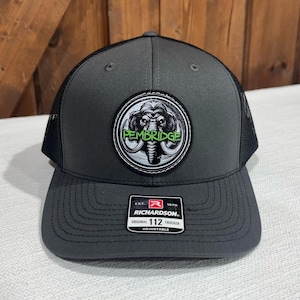 May include: A gray and black trucker hat with a black and white patch featuring a mammoth and the text "PEMBRIDGE" in green. The hat is adjustable and has the Richardson brand logo.