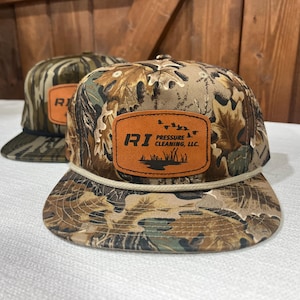 May include: Two camouflage baseball caps with a flat brim and a brown leather patch. The patch on the front cap has the letters "RI" and the words "Pressure Cleaning, LLC" with a graphic of ducks and reeds. The caps have a rope detail around the brim.