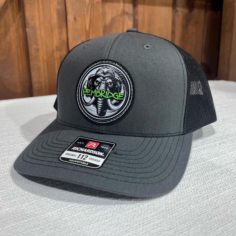 May include: A gray and black trucker hat with a black and white patch featuring a mammoth and the text "PEMBRIDGE". The patch has a green outline. The hat is adjustable and has the Richardson 112 logo.