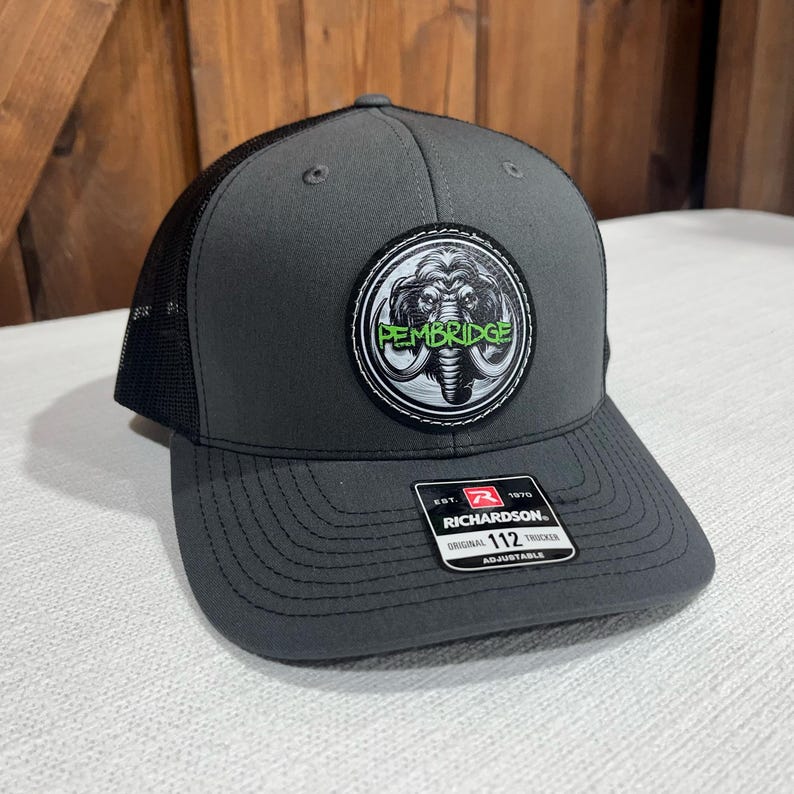 May include: A gray and black trucker hat with a green and white patch featuring a mammoth and the text "Pembridge". The hat is adjustable and has the Richardson logo on the back.