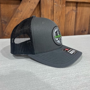 May include: A gray and black trucker hat with a black mesh back. The hat has a circular patch with a white elephant design and the text "PEMBROKE" in green.