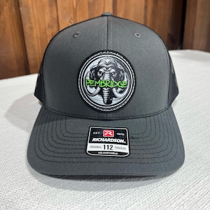 May include: A gray and black trucker hat with a black and white mammoth graphic and the text "Pembridge" in green. The hat is made by Richardson and has an adjustable strap.
