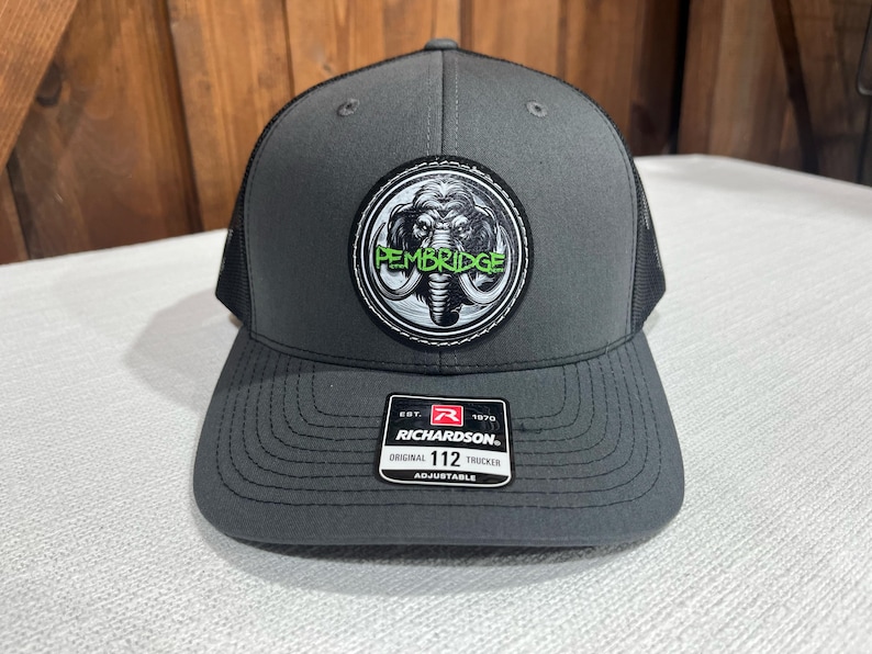 May include: A gray and black trucker hat with a black and white mammoth graphic and the text "Pembridge" in green. The hat is made by Richardson and has an adjustable strap.