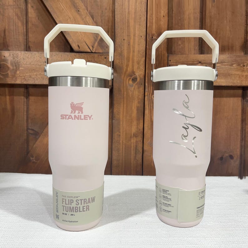 May include: Two pink stainless steel tumblers with white lids and handles. The tumbler on the left has the Stanley logo and the text "The Iceflow Flip Straw Tumbler 20 oz / 591 mL Active Hydration". The tumbler on the right has the name "Layla" written on it.