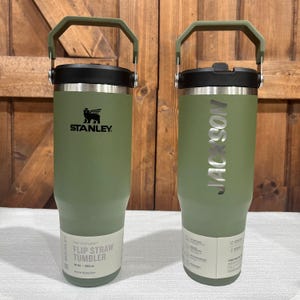 May include: Two green stainless steel tumblers with handles. The tumbler on the left has the Stanley logo and the text "The  Flip Straw Tumbler 30 oz | 890 ml Active Hydration". The tumbler on the right has the text "JACKSON" engraved on the side.