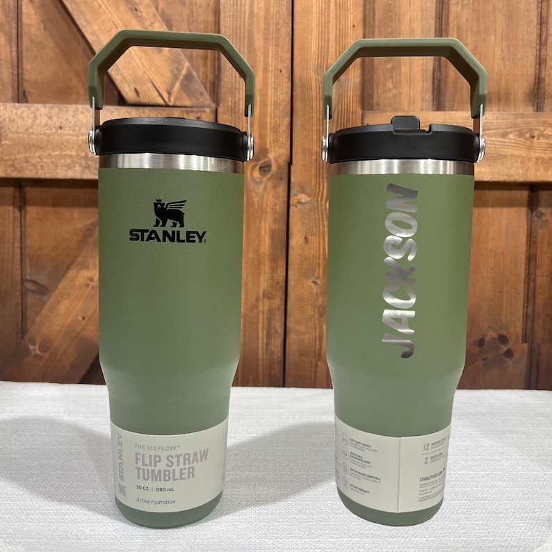 May include: Two green stainless steel tumblers with handles. The tumbler on the left has the Stanley logo and the text "The  Flip Straw Tumbler 30 oz | 890 ml Active Hydration". The tumbler on the right has the text "JACKSON" engraved on the side.