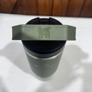 May include: A green stainless steel travel mug with a black lid and a handle. The lid has a logo of a winged creature with a crown on its head.
