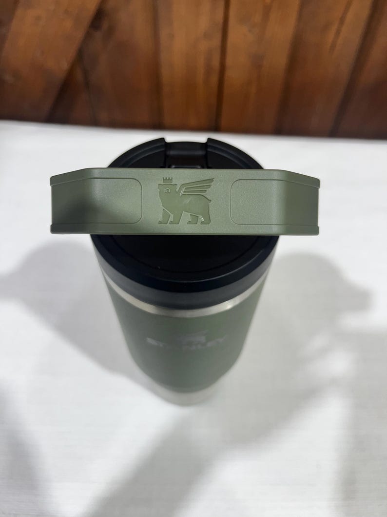 May include: A green stainless steel travel mug with a black lid and a handle. The lid has a logo of a winged creature with a crown on its head.
