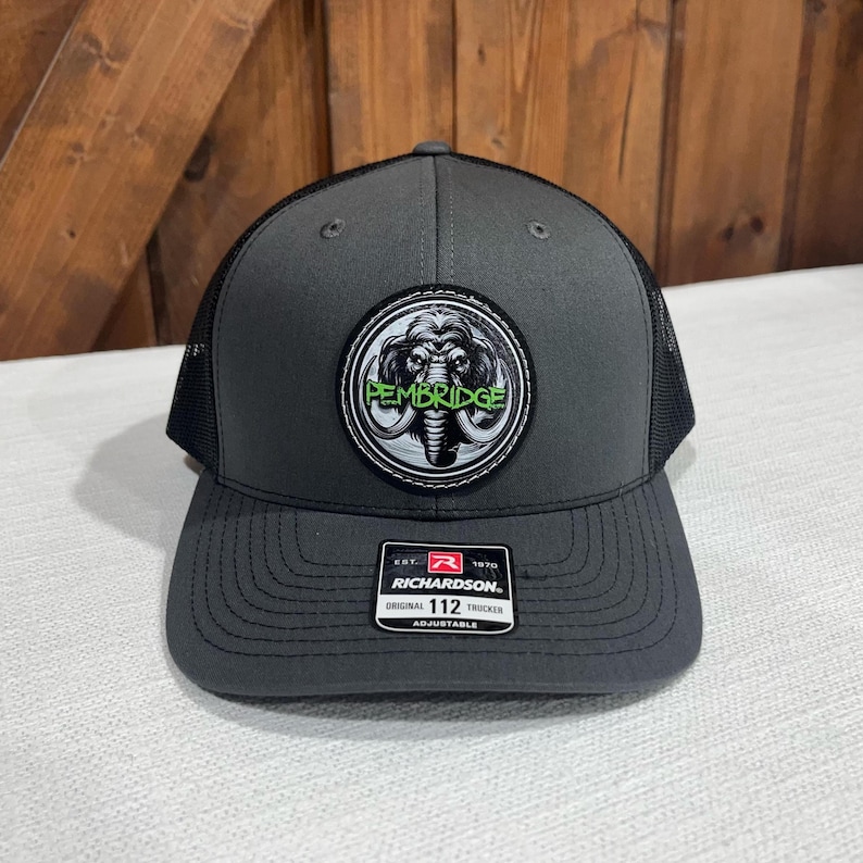 May include: A gray and black trucker hat with a black and white patch featuring a mammoth and the text "PEMBRIDGE" in green. The hat is adjustable and has the Richardson brand logo.