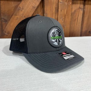May include: A gray and black trucker hat with a black mesh back. The hat has a round patch with a white mammoth and the text "PEMBRIDGE" in green.