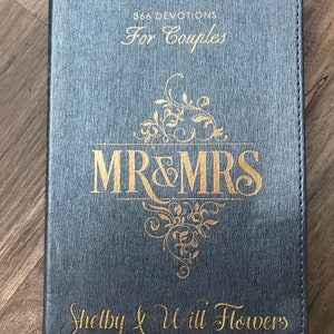 May include: A gray leather-bound journal with gold lettering. The cover reads "366 Devotions For Couples" and features a gold design with the words "Mr & Mrs" and the names "Shelby & Will Flowers".